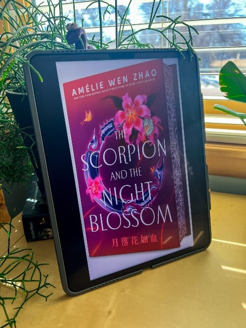 The Scorpion and the Night Blossom by Amélie Wen Zhao is displayed on an iPad leaning against a black planter