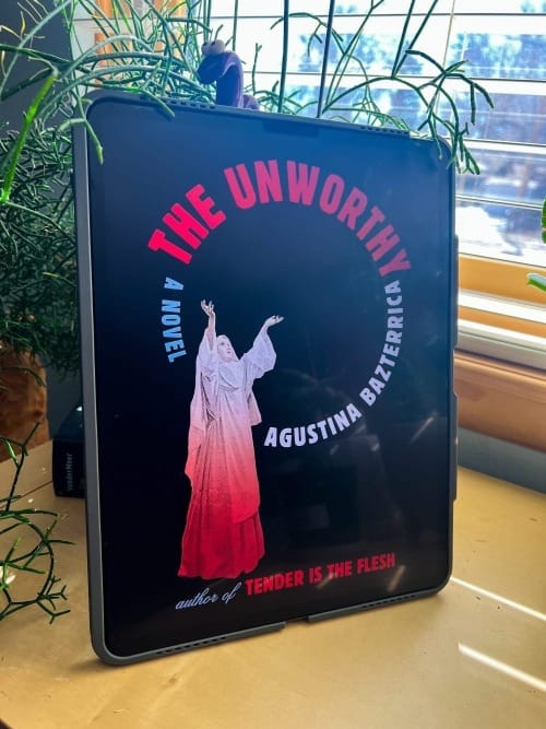 Cover of the Unworthy by Agustina Bazterrica is displayed on an iPad leaning against a black planter